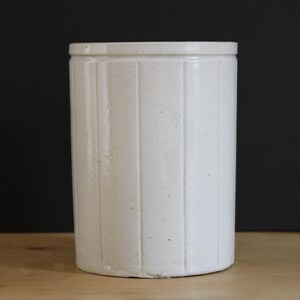 May include: A cylindrical off-white ceramic crock featuring a series of vertical recessed panels around its exterior. The vessel has a smooth, glazed finish and a flat, reinforced rim at the top, presenting a simple, functional, and sturdy design.