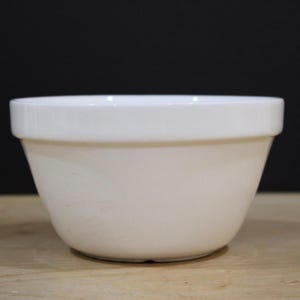 Może przedstawiać: Jednolita biała ceramiczna miska do mieszania o gładkim, błyszczącym wykończeniu. Misa posiada szeroki, płaski brzeg u góry oraz głęboki, zaokrąglony kształt, który zwęża się ku mniejszej, okrągłej podstawie, tworząc czysty i prosty projekt.