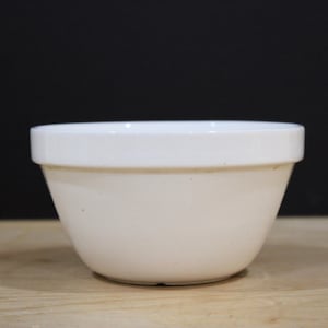 May include: A solid white ceramic mixing bowl with a smooth, glossy finish and a wide, flat rim. The bowl features a deep, rounded basin that tapers toward a smaller circular base, presenting a clean, functional design for kitchen use.