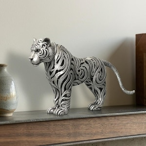 May include: A silver-colored tiger figurine featuring an intricate, openwork filigree pattern across its body. The sculpture depicts the animal in a standing position with its tail extended, showcasing detailed metallic textures and curved lines.