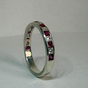 May include: A silver-toned band ring featuring an alternating channel-set pattern of square-cut deep red rubies and clear white stones. The metal band has a smooth, polished finish and a consistent width throughout its circular design.