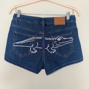 May include: Dark blue denim shorts featuring a white line art illustration of an alligator spanning across both back pockets. The shorts include a tan leather-like patch on the waistband and are displayed on a light-colored plastic clothes hanger.