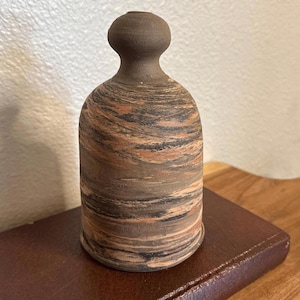 May include: A cylindrical ceramic vessel with a narrow, rounded neck and a wide base. The body features a marbled pattern of earthy brown, terracotta, and black tones, while the neck is a solid, matte dark brown finish with a small opening.