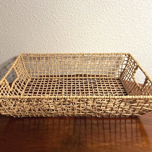 May include: A rectangular, open-weave basket crafted from light tan, natural fibers. The basket features a grid-like pattern throughout its base and sides, with integrated handles on two opposing ends, providing a structured, shallow storage tray.