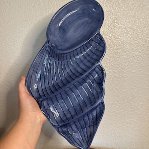 May include: A large, deep blue ceramic serving dish shaped like a conch shell. The dish features a glossy finish with visible brushstroke textures and distinct, curved ridges that divide the interior into several sections for food presentation.