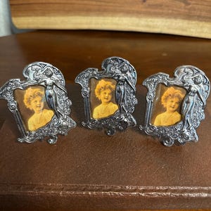 May include: Three small silver-toned metal picture frames with an ornate, embossed design featuring a standing figure on the right side. Each frame holds a miniature portrait of a young child with curly blonde hair wearing a yellow shirt.