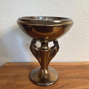 May include: A metallic bronze-colored pedestal bowl featuring a wide, circular rim and a central stem supported by two symmetrical, wing-like decorative elements that connect the base to the bowl, creating a cohesive, reflective finish.