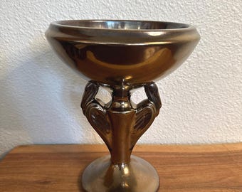 May include: A metallic bronze-colored pedestal bowl featuring a wide, circular rim and a central stem supported by two symmetrical, wing-like decorative elements that connect the base to the bowl, creating a cohesive, reflective finish.