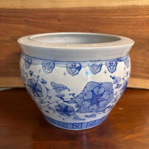May include: A round ceramic planter featuring a white base with blue floral patterns and vine motifs. The rim is decorated with a geometric Greek key border. The exterior surface displays hand-painted blue flowers and leaves.