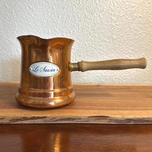 May include: A copper sauce pot with a flared rim and a long, light-colored wooden handle. The body of the pot features an oval white label with a blue border and the text Le Saucier printed in blue script. The metal surface has a polished finish.