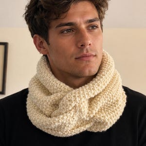 Puede incluir: Una bufanda infinita de punto grueso en color crema blanquecino. La bufanda presenta un patrón de punto bobo grueso y texturizado, envuelta en un doble bucle alrededor del cuello, sobre una camiseta negra lisa de manga larga.