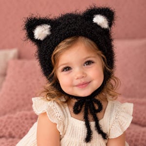 May include: A black fuzzy knit bonnet featuring two pointed cat-like ears with white centres. The bonnet is constructed from a textured, curly yarn and includes two long black ties that fasten under the chin to secure the head covering in place.