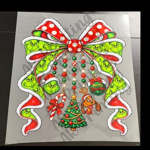 Peut inclure: Un grand nœud rouge à pois blancs avec un motif vert Grinch, agrémenté de perles suspendues, d'un sucre d'orge, d'un sapin de Noël décoré, d'un chien marron avec un bonnet de Père Noël et d'une boule verte avec le texte Merry Grinchmas.