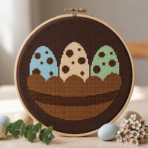 May include: A circular wooden embroidery hoop containing a cross-stitch pattern of three speckled eggs in light blue, cream, and mint green, nestled inside a dark brown basket against a solid dark brown background with a tight grid texture.