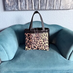 May include: A small tote bag featuring a leopard print pattern on the front panel, complemented by a black glittery texture on the upper rim and handles. The bag has a structured rectangular shape with two top handles for carrying.