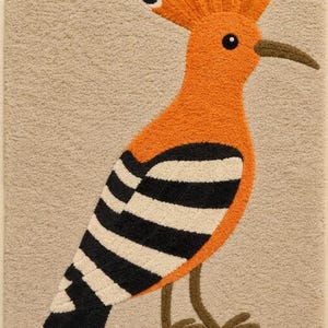 Op de afbeelding: Een rechthoekig getuft tapijt met een afbeelding van een hop. De vogel heeft een feloranje lichaam, een opvallende kuif met zwart getipte veren en zwart-wit gestreepte vleugels. Hij staat op dunne bruine poten tegen een beige achtergrond.
