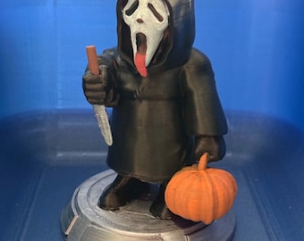 May include: A 3D printed figurine of a character wearing a black hooded robe and a white ghost mask with a red tongue sticking out. The figure holds a silver-colored knife in one hand and an orange pumpkin in the other, standing on a circular base.
