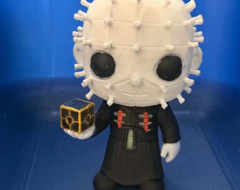 May include: A stylized figurine featuring a white head covered in protruding spikes, large black circular eyes, and a black robe with red accents. The figure holds a small black cube with gold-colored geometric lines and markings in one hand.