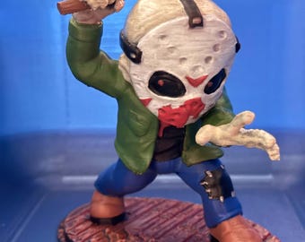 May include: A stylized figurine of a character wearing a white hockey mask with red markings, a green jacket, and blue jeans. The figure holds a yellow axe with red paint resembling blood and stands on a textured, reddish-brown oval base.