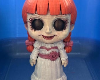 May include: A stylized doll figure with bright orange hair styled in bangs and two side braids. The doll has large black eyes, a pale face, and a wide, thin-lipped smile. It wears a white dress with a pink waistband featuring a central flower detail.