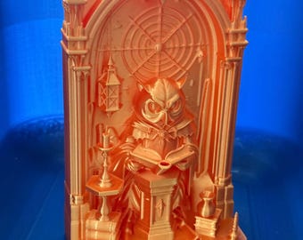 May include: A 3D printed orange figurine featuring an owl wearing robes and reading a book while seated at a pedestal. The scene is set within an arched architectural frame with a spiderweb design, a hanging lantern, and stacks of books.