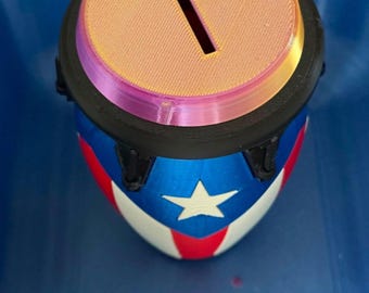 May include: A cylindrical coin bank featuring a top lid with a central rectangular slot. The body is decorated with a blue, white, and red pattern resembling a flag, including a prominent white star. The lid has a gradient of peach and pink tones.