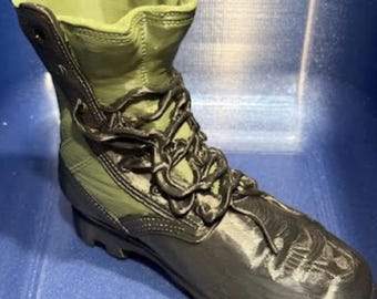 May include: A 3D printed combat boot sculpture featuring an olive green upper section and a black lower section with black laces. The boot has a high-top design with a visible eyelet at the top collar and a textured, layered surface finish.