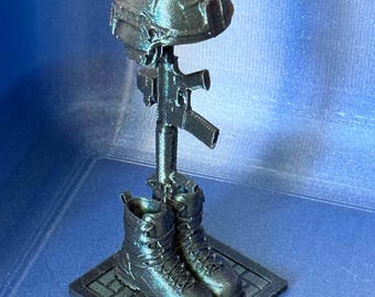 May include: A 3D printed memorial display featuring a black tactical helmet resting atop a rifle, which is positioned vertically between a pair of black combat boots. The entire assembly is mounted on a square base with a brick-like pattern.