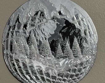 May include: A circular silver-colored ornament featuring a 3D-printed relief of a winter forest scene with pine trees and mountains. The design has a textured, layered appearance with a dark background visible through the cutouts and a hanging loop.