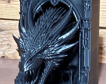 May include: A matte black 3D printed bookend featuring a highly detailed dragon head emerging from a gothic archway. The dragon has sharp scales, prominent horns, and an open mouth with visible teeth, set against a rectangular frame structure.