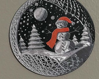 May include: A circular ornament featuring a black background with a silver 3D-printed winter scene. The design includes a snowman wearing a bright orange scarf and hat, surrounded by silver pine trees, a moon, and stars, with a textured lattice border.