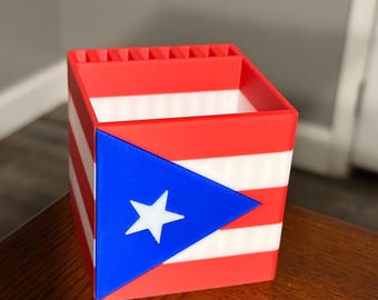 May include: A square desk organizer featuring the design of the Puerto Rican flag with horizontal red and white stripes and a blue triangle on the front containing a single white star. The top edge includes a series of small vertical slots.