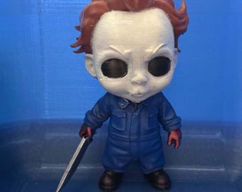 May include: A stylized figurine featuring a character with a white mask, dark hollow eyes, and messy brown hair. The figure wears a blue jumpsuit and holds a silver-colored knife in one hand. The design has a large head and small body proportions.