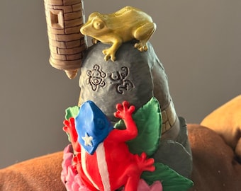 May include: A decorative figurine featuring a gray stone-like structure with two carved symbols, a tan watchtower, a gold-colored frog on top, and a red, white, and blue frog with a star emblem positioned among pink flowers and green leaves.