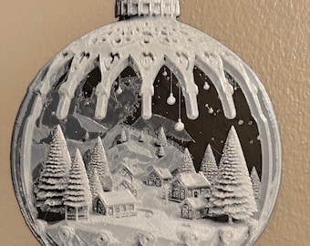 May include: A circular, flat, white and black ornament featuring a detailed winter village scene with snow-covered houses and pine trees. The design is framed by an arched structure resembling a bauble top, hanging from a metal nail.