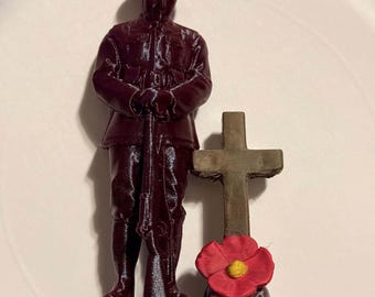 May include: A dark maroon 3D-printed figurine of a soldier in a helmet standing with a rifle. Beside the soldier is a small tan cross with a bright red poppy flower featuring a yellow center at the base, all mounted on a rectangular platform.