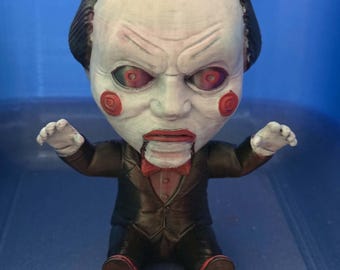May include: A stylized figurine of a ventriloquist dummy with a large head, pale white skin, and dark hair. The figure has red spiral cheeks, red lips, and red eyes. It wears a black suit jacket, a red bow tie, and red shoes, with arms outstretched.