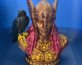 May include: A bronze-colored bust of a figure wearing a winged helmet and ornate armor with yellow accents. The figure has long magenta hair and a black bird with yellow talons perched on the right shoulder. The base features carved runic text.