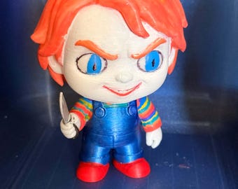 May include: A stylized figurine featuring a character with bright orange hair, pale skin, and piercing blue eyes. The figure wears blue overalls over a multi-colored striped long-sleeve shirt and red shoes, while holding a small silver-colored knife.