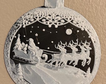 May include: A circular Christmas ornament featuring a white 3D-printed relief of Santa Claus in a sleigh pulled by reindeer against a black background. The design includes a starry night sky, a moon, and decorative lace-like borders at the top and bottom.