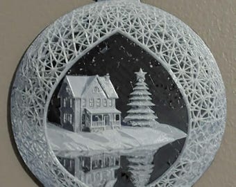 May include: A circular white 3D printed ornament featuring a detailed winter scene with a house and a Christmas tree. The outer border has a complex geometric lattice pattern. The central scene is set against a dark background, creating a high-contrast visual effect with a reflective surface below the house and tree.