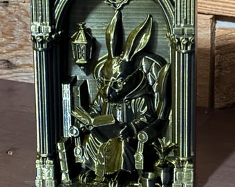 May include: A three-dimensional rectangular wall plaque featuring a metallic gold and black finish. The relief depicts a rabbit wearing glasses and a robe, sitting in a chair while reading a book inside a gothic archway with a lantern.