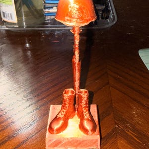May include: A copper-colored 3D printed memorial statue featuring a military helmet resting on top of a rifle, which is positioned vertically between a pair of combat boots. The entire assembly is mounted on a square base of the same color.