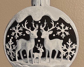 May include: A circular white and black ornament featuring a raised relief design of two adult deer and one fawn standing in a snowy landscape. The scene is framed by white snowflakes against a black background, with a hanging loop at the top.