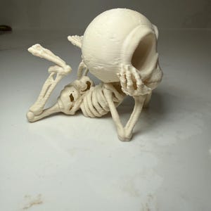 May include: An off-white, 3D-printed figurine of a skeleton in a reclining pose. The skeleton features a large, rounded cranium, visible rib cage, and articulated limbs, with one hand resting near the face and legs bent at the knees.