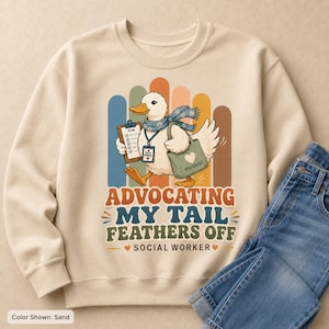 May include: A sand-colored crewneck sweatshirt featuring a graphic of a white duck wearing a blue patterned scarf and a lanyard. The duck holds a clipboard labeled PLAN and a green bag labeled RESOURCES. Text reads ADVOCATING MY TAIL FEATHERS OFF SOCIAL WORKER.
