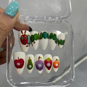 May include: A set of ten almond-shaped press-on nails with a white base. The top row features a painted green caterpillar with a red head. The bottom row displays various painted fruits including a red apple, green pear, purple plum, red strawberry, and orange.