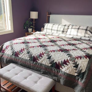 May include: A bedroom featuring a bed with a white, burgundy, and dark green geometric patterned quilt, plaid pillows, and a light gray upholstered headboard. A light gray tufted bench with a wooden base sits at the foot of the bed.
