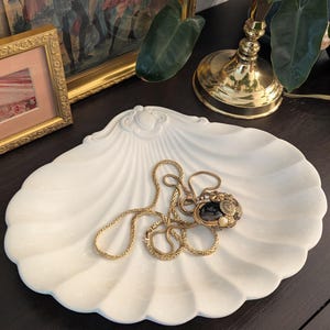 May include: A white ceramic dish shaped like a large scallop shell with a decorative embossed detail at the top. Resting inside the shell dish is a gold-toned metal necklace featuring a dark oval pendant surrounded by small metallic accents.