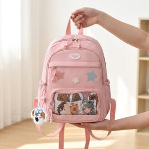 May include: A light pink backpack featuring a top handle, adjustable shoulder straps, and a front zippered pocket with a clear display window. The bag is decorated with three star-shaped patches and a plush dog keychain attached to the side zipper.